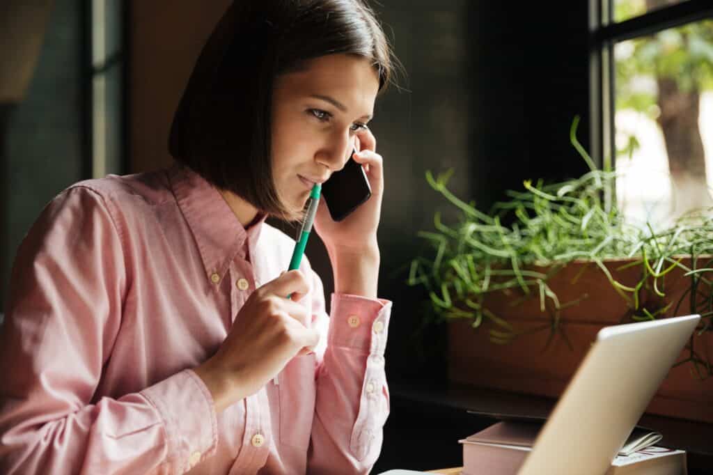 A woman holding a pen in front of the computer is speaking with the moving company on the phone